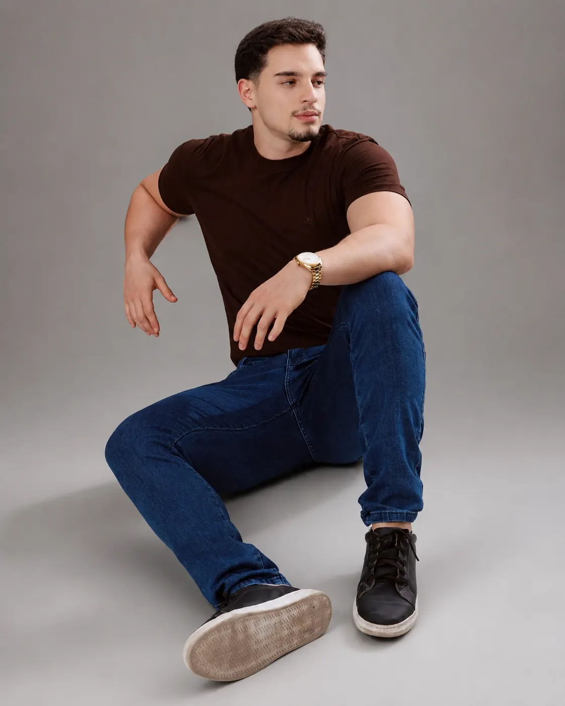 Modelo masculino sentado com camiseta marrom e calça jeans